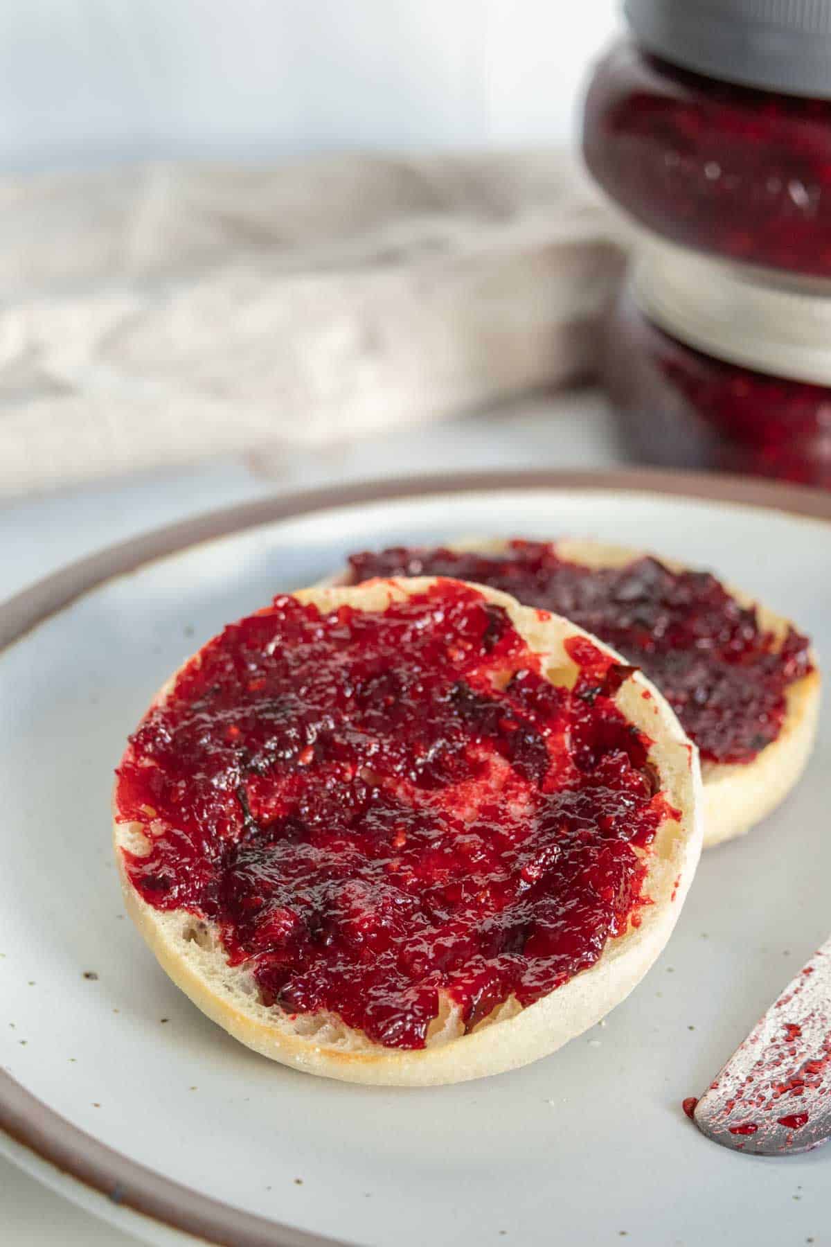 A round piece of bread topped with a spread of red jam on a white plate, with jars of the same jam in the background.