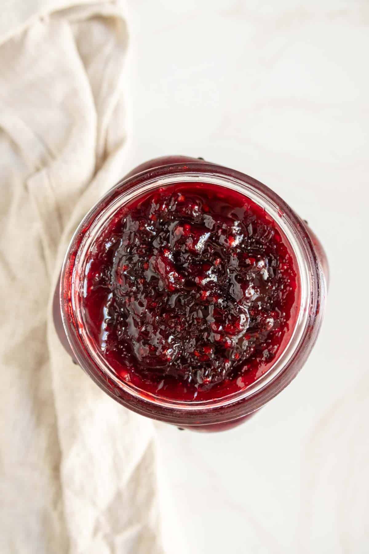 Top view of a glass jar filled with a chunky dark red preserve, likely berry jam, placed on a light-colored surface with a beige cloth nearby.