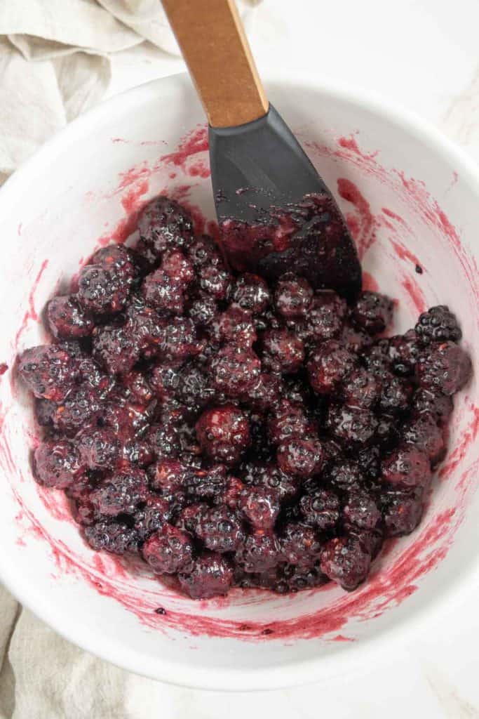 A bowl filled with mashed blackberries mixed with a dark, viscous liquid. A black spatula with a wooden handle rests on the edge of the bowl.