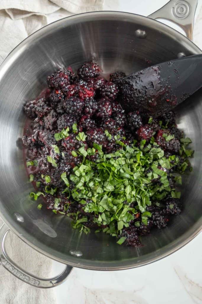 A stainless steel pot containing crushed blackberries and chopped fresh herbs, with a black spatula partly visible inside the pot.