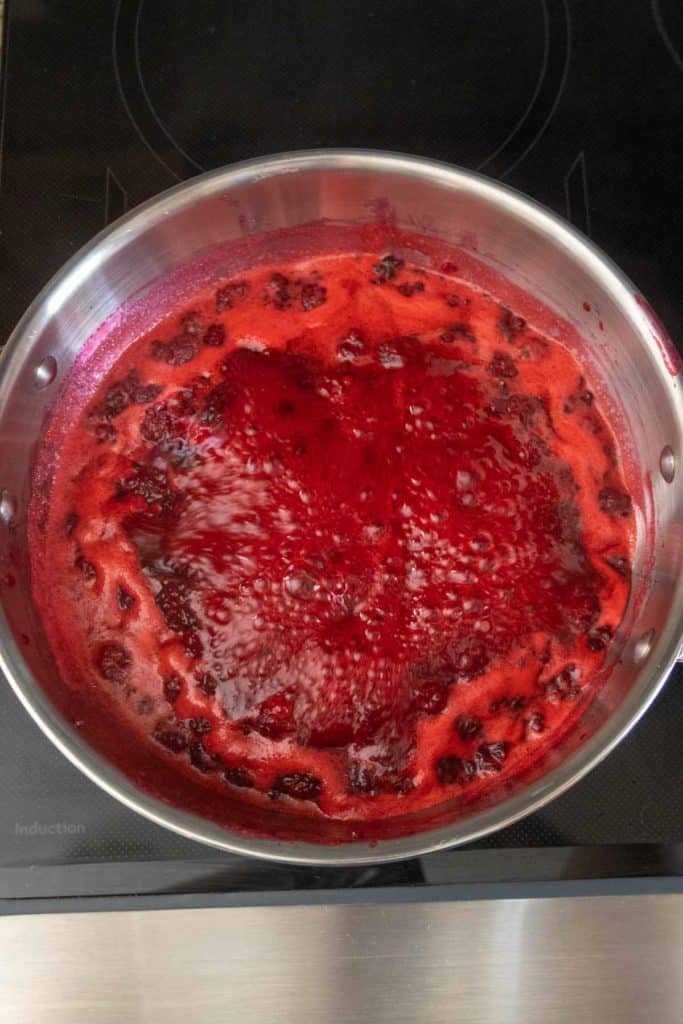 A stainless steel pot on an induction stove with bubbling, simmering red liquid, possibly a berry reduction or sauce, with visible chunks of fruit.