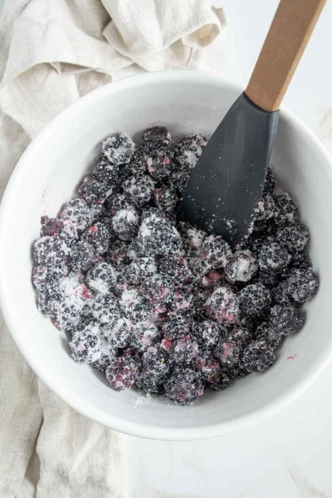 A white bowl filled with blackberries coated in sugar, with a black spatula resting inside. A white cloth is visible in the background.