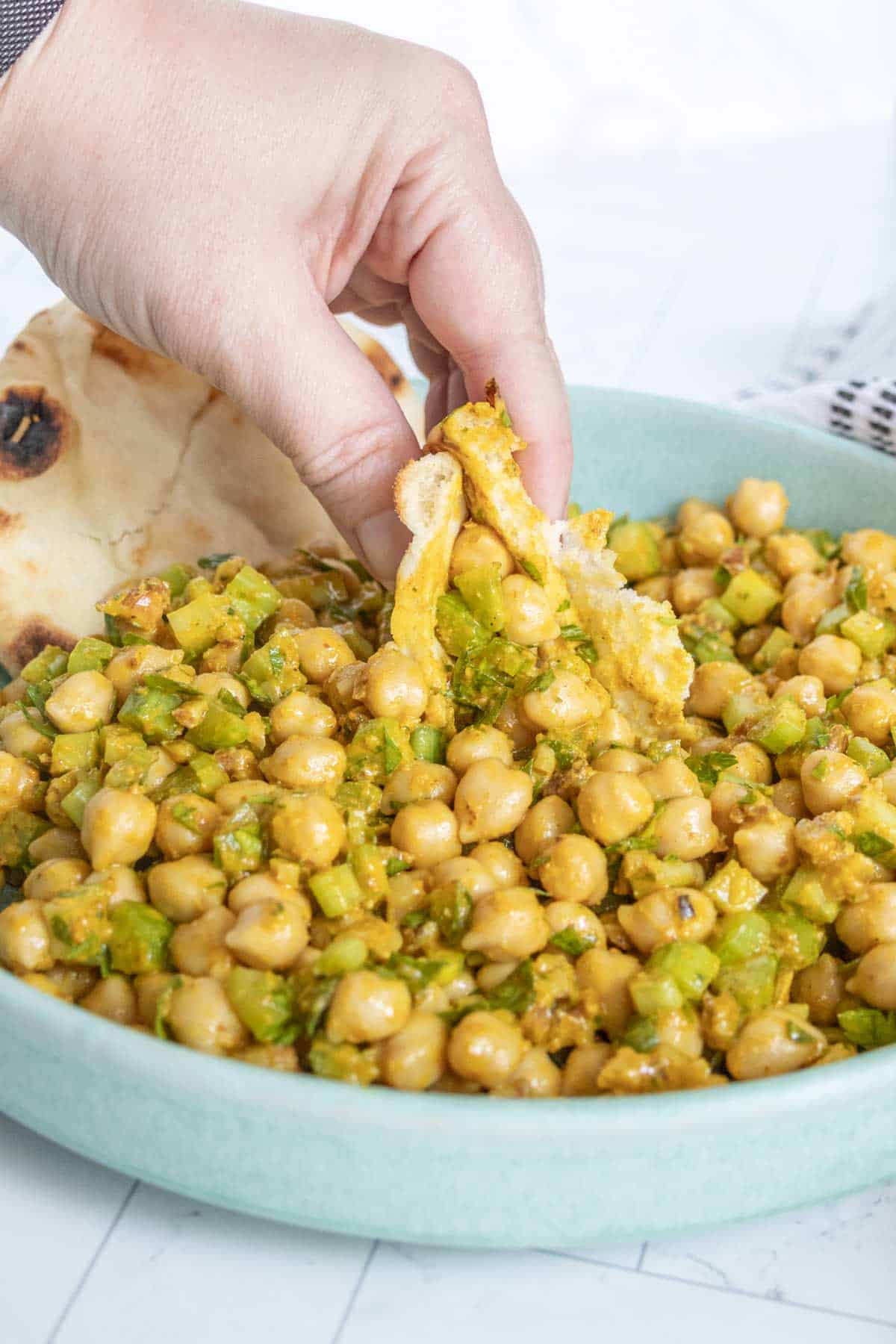 A hand holding a piece of bread is dipping it into a dish of chickpeas mixed with diced vegetables, served in a light blue bowl.