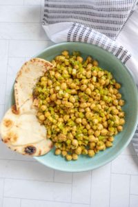 A green bowl filled with chickpea salad containing diced vegetables, accompanied by two pieces of naan bread on the side. A white and black patterned cloth napkin is partially visible.