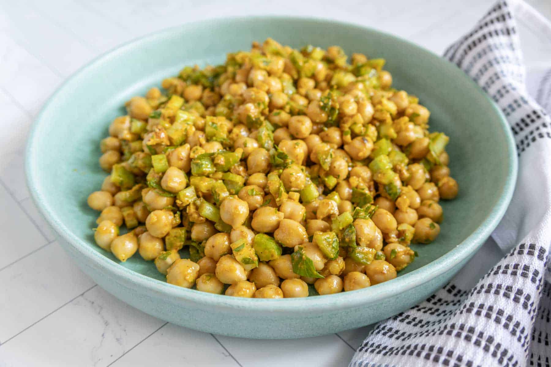 A bowl filled with a chickpea and diced green vegetable salad, placed on a tiled surface next to a folded white and black striped cloth.