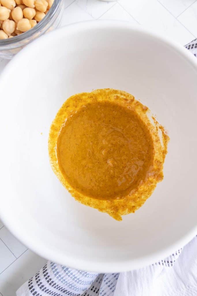 A white bowl containing a partially mixed orange-brown paste is on a counter next to a jar of chickpeas and a striped cloth.