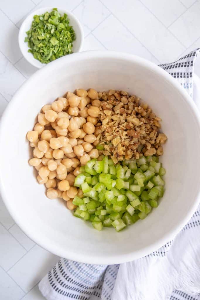 A white bowl contains chickpeas, chopped walnuts, and diced celery. A small bowl with chopped herbs is in the background, with a striped towel nearby.