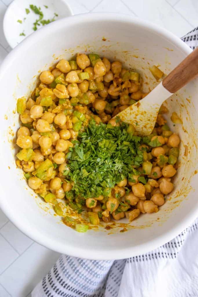 A white bowl contains a mixture of chickpeas, chopped herbs, and celery with a wooden spoon, set on a striped cloth.