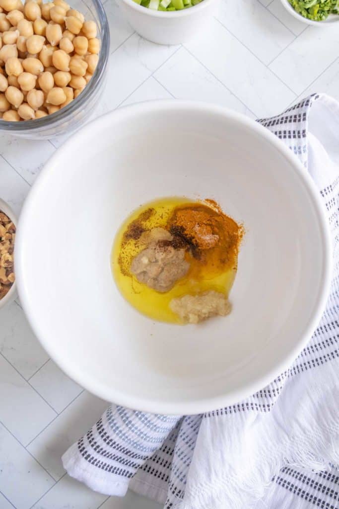 A large white bowl contains oil, mashed chickpeas, and spices. Surrounding it are bowls with chickpeas, chopped vegetables, and nuts on a white tiled surface with a striped towel.