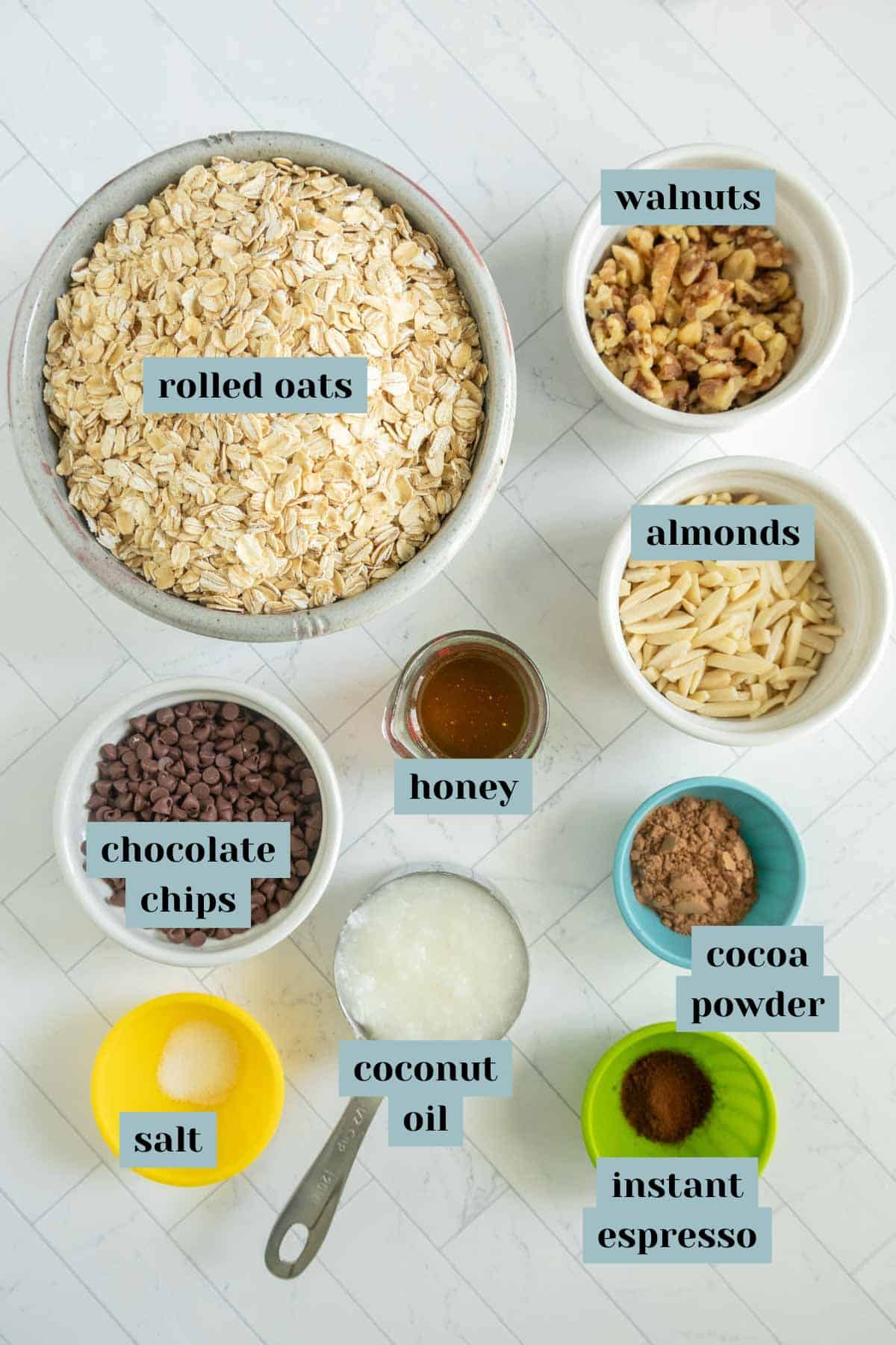 A flat lay of various ingredients including rolled oats, walnuts, almonds, honey, chocolate chips, cocoa powder, instant espresso, salt, and coconut oil, all in separate bowls on a white surface.