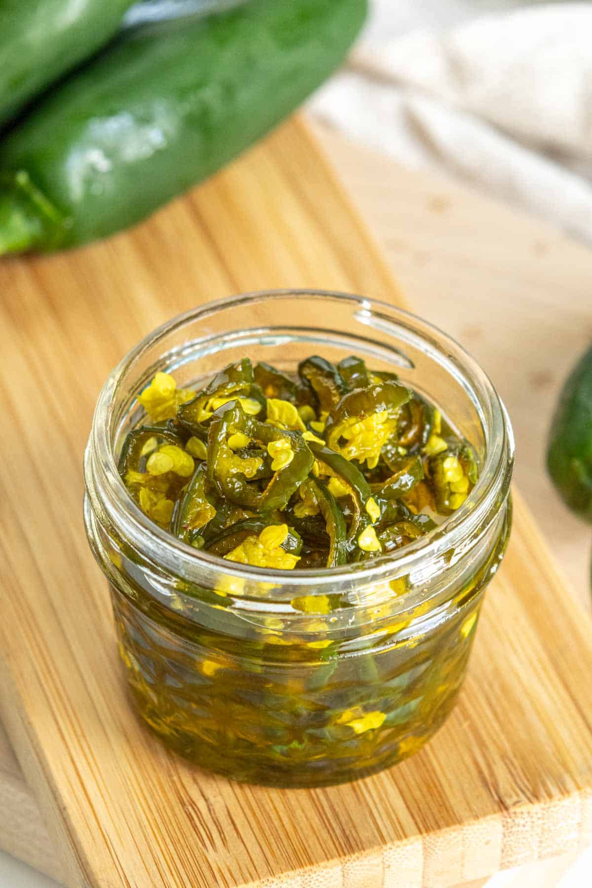 A small glass jar filled with sliced candied jalapeños, placed on a wooden surface with whole jalapeños in the background.