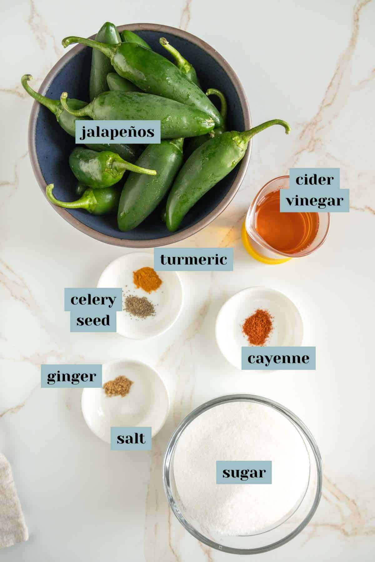 Ingredients for candied jalapeños include fresh jalapeños, cider vinegar, turmeric, cayenne pepper, celery seed, ground ginger, salt, and sugar, all arranged on a marble surface.