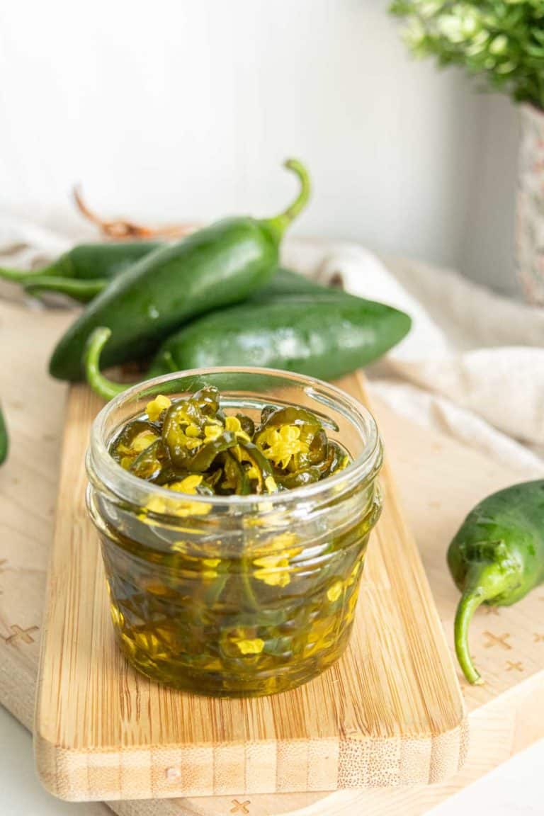 A small glass jar filled with pickled jalapeño slices is placed on a wooden cutting board with whole jalapeños scattered around.