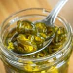 A spoon raises sliced candied jalapeños from a glass jar, placed on a wooden surface.