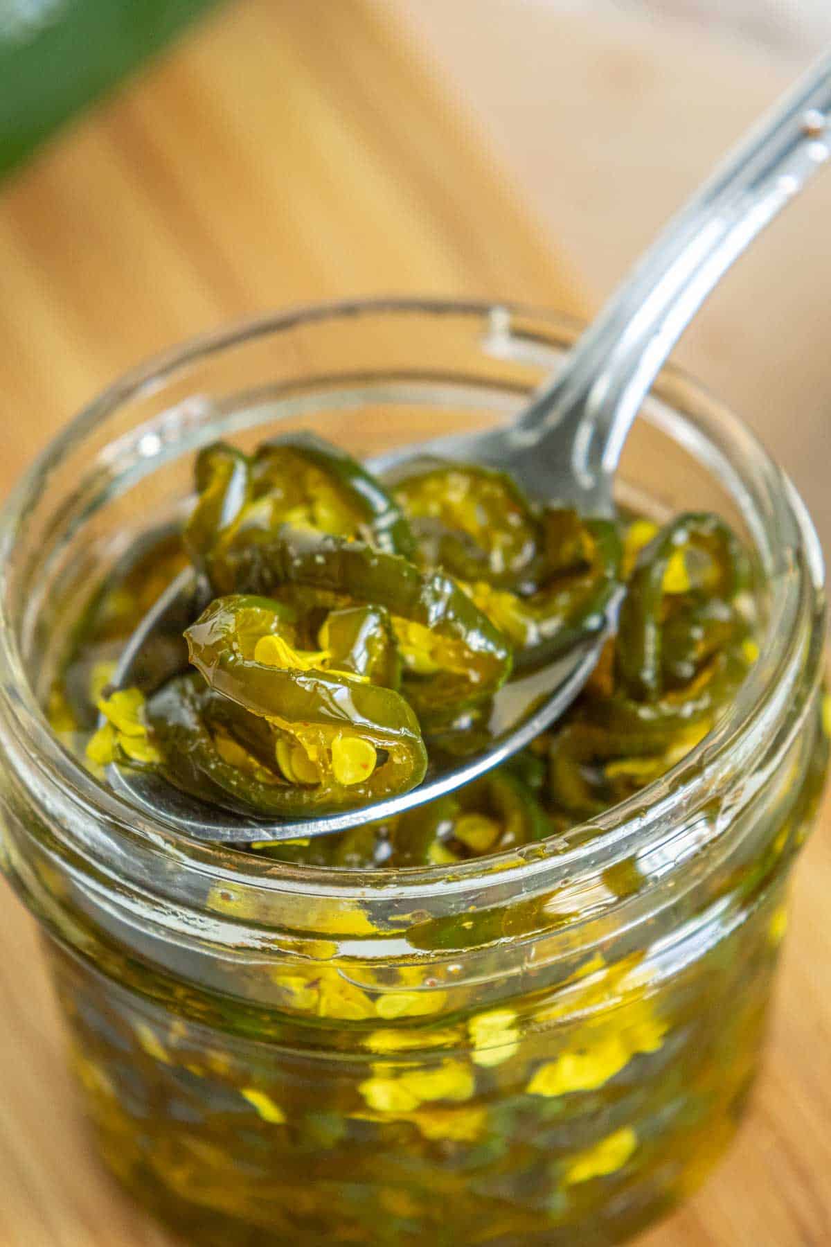 A spoon raises sliced candied jalapeños from a glass jar, placed on a wooden surface.