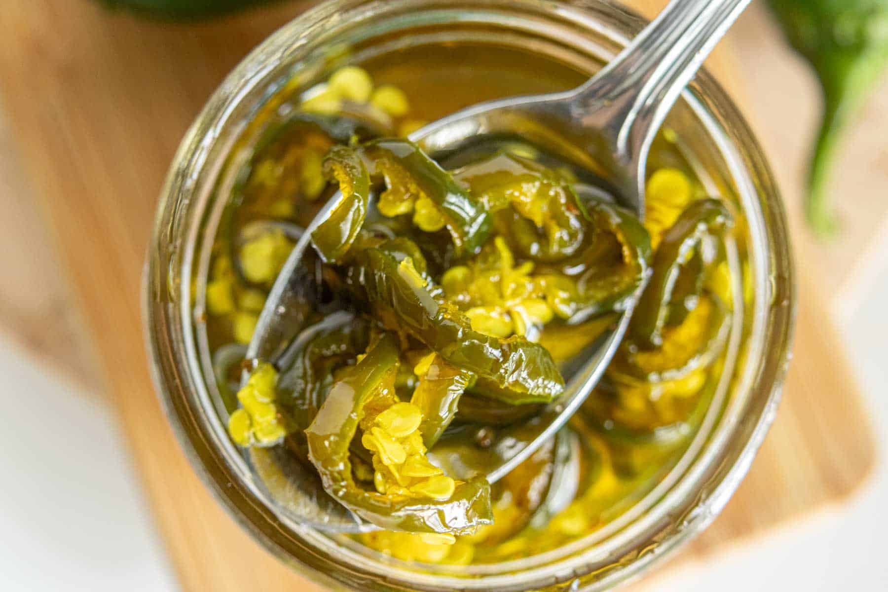 A close-up shot of a jar filled with candied jalapeños, with a spoon lifting some slices out. The jalapeños are green and submerged in a liquid.