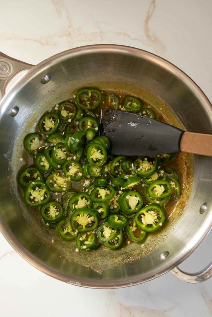 A metal pot contains sliced green jalapeños in a liquid with a spatula placed inside.