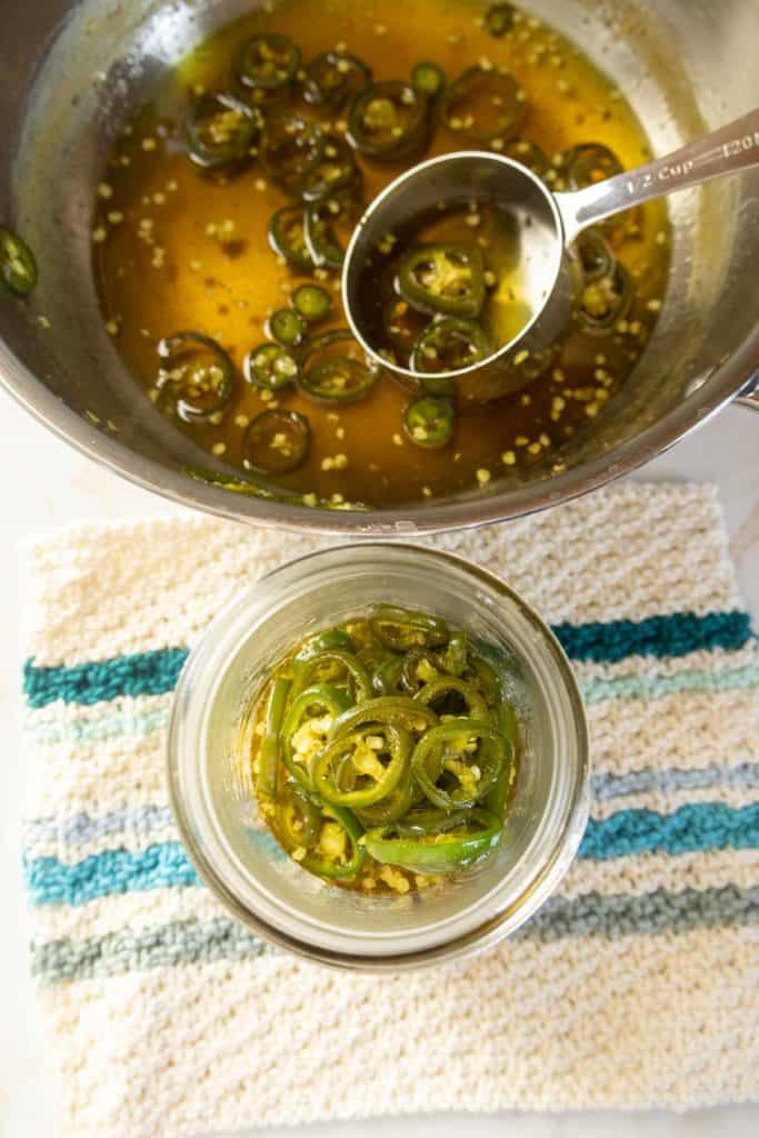 A jar being filled with sliced jalapeños and pickling liquid using a ladle. The process takes place on a striped cloth surface.