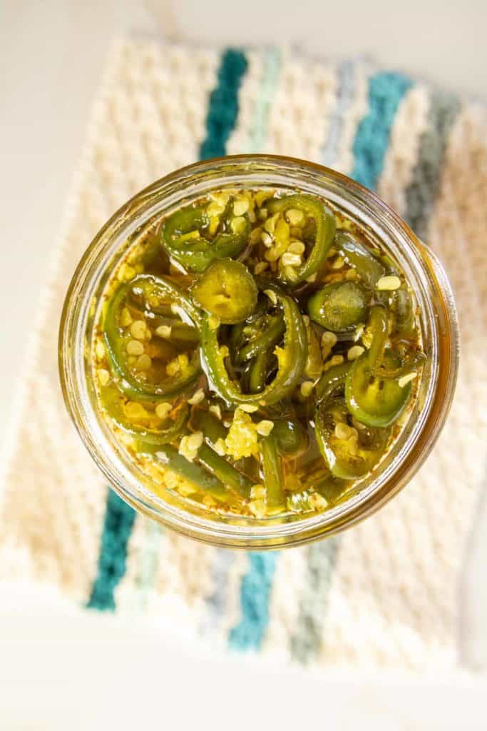 A top-down view of a jar filled with sliced candied jalapeños on a textured cloth with blue and beige stripes.