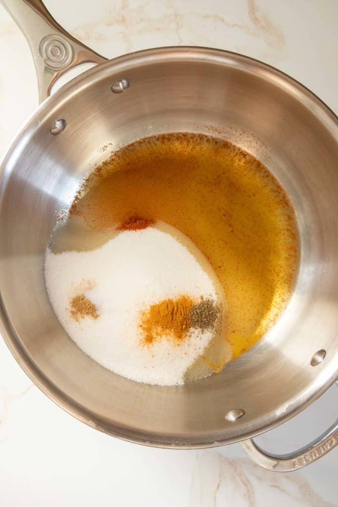 A stainless steel pot on a marble countertop contains oil, sugar, and brown seasoning.