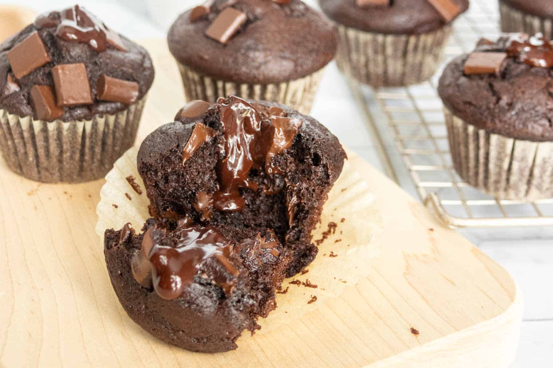 A broken chocolate muffin with melted chocolate on a wooden board, surrounded by whole chocolate muffins, some on a cooling rack in the background.