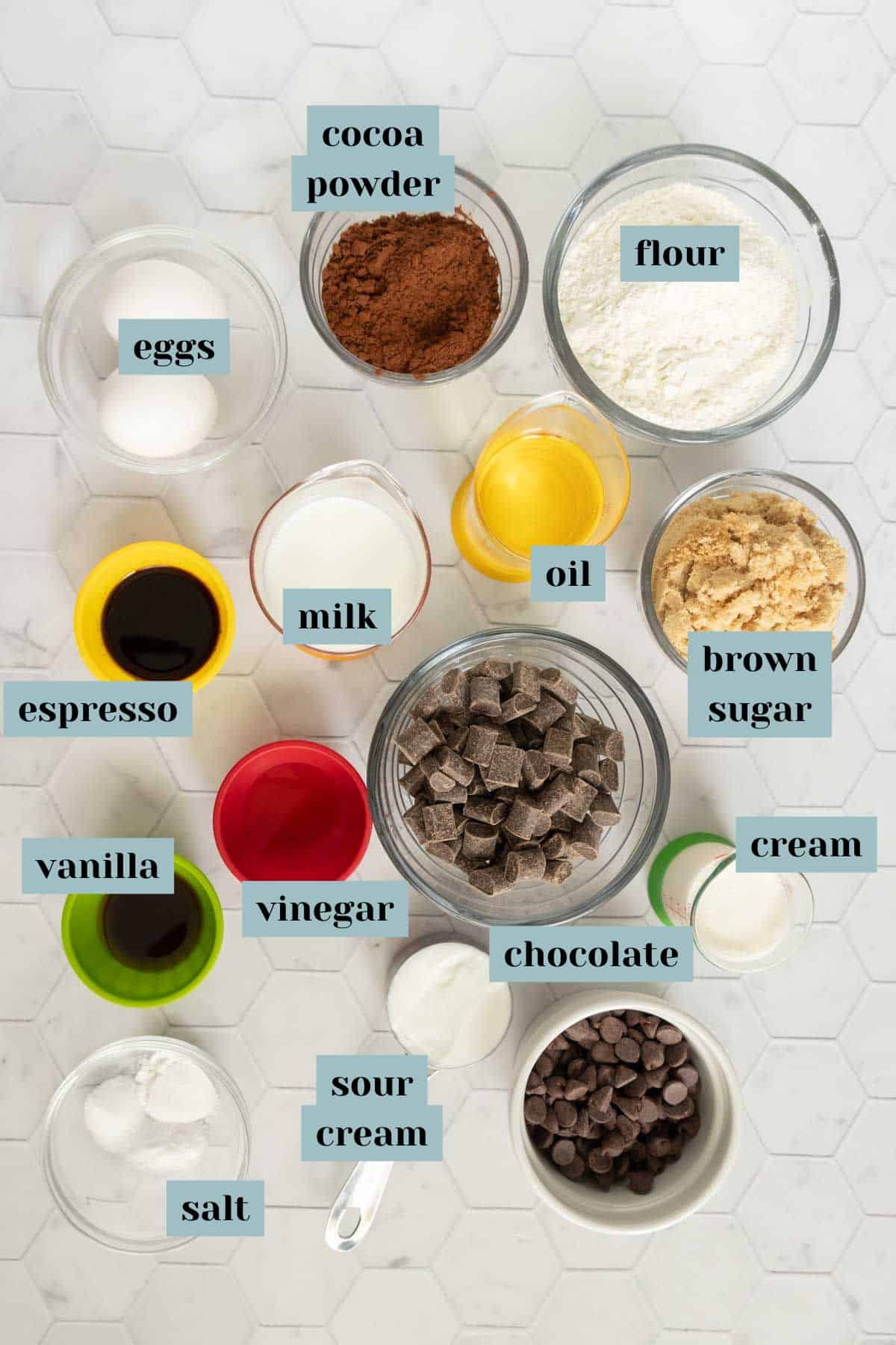 A photo of various baking ingredients in bowls and cups, including flour, eggs, cocoa powder, chocolate, brown sugar, milk, cream, sour cream, oil, vinegar, espresso, vanilla, and salt.