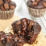 A close-up of a chocolate muffin, broken in half to show a gooey chocolate center, surrounded by more muffins and a small bowl of chocolate chunks in the background.