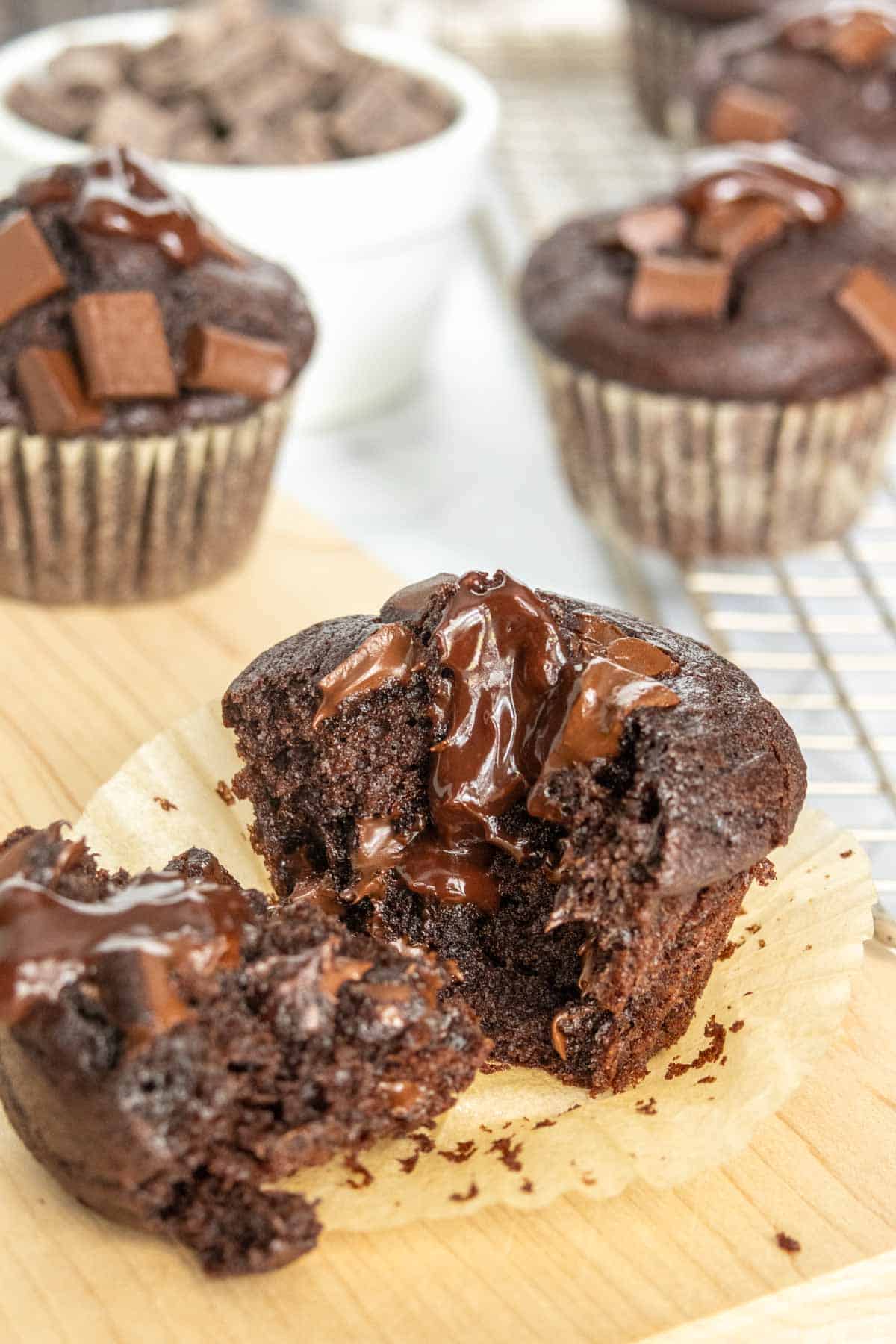 A close-up of a chocolate muffin, broken in half to show a gooey chocolate center, surrounded by more muffins and a small bowl of chocolate chunks in the background.