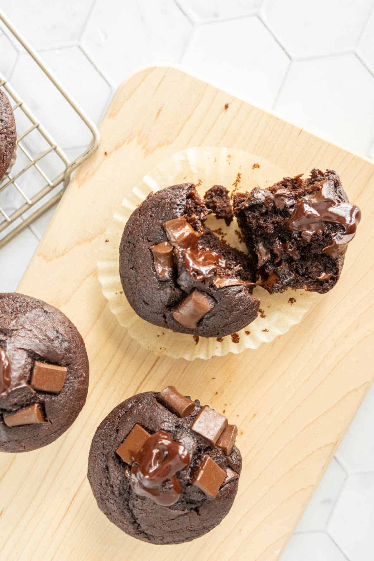 Chocolate muffins on a wooden surface, some topped with melted chocolate chunks. One muffin is partially unwrapped and broken in half, revealing a moist interior. A cooling rack is partially visible.