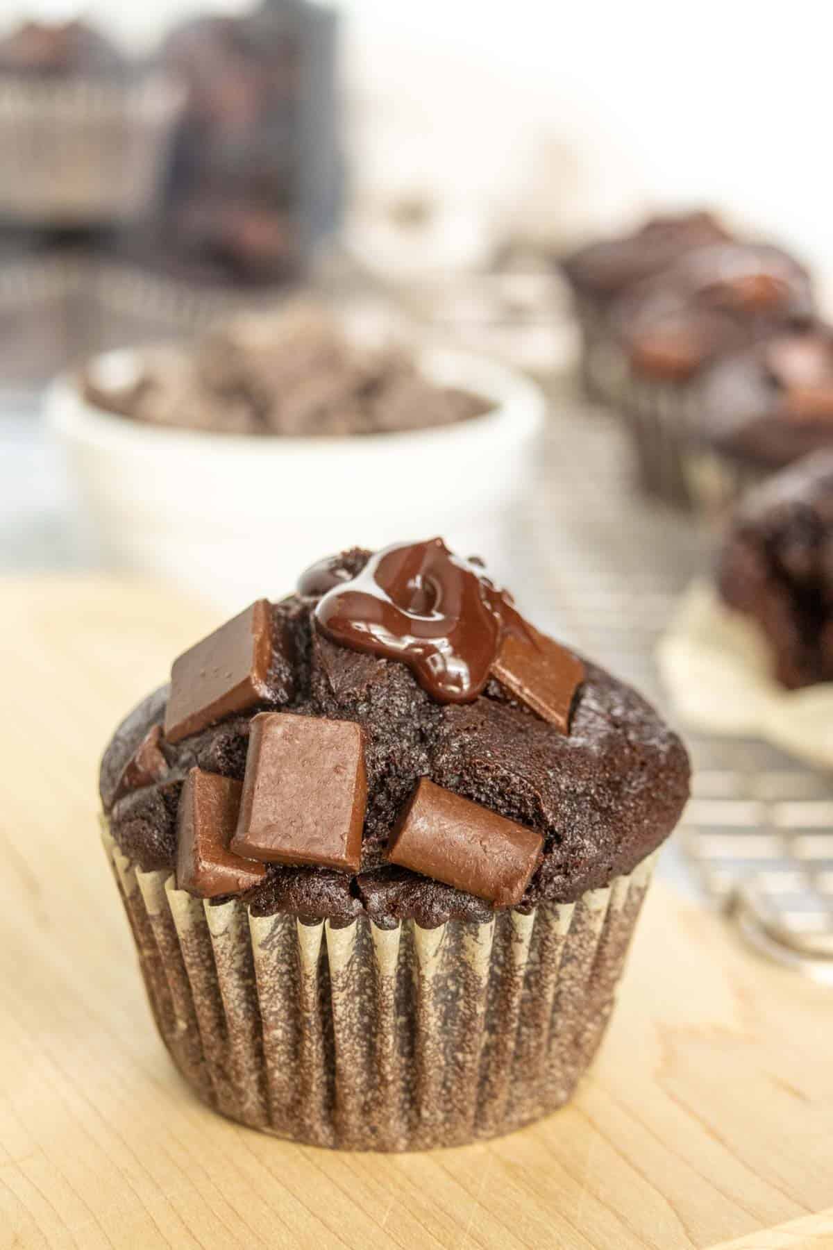 Close-up of a chocolate muffin topped with chocolate chunks and a glossy drizzle, displayed on a wooden surface with more muffins blurred in the background.