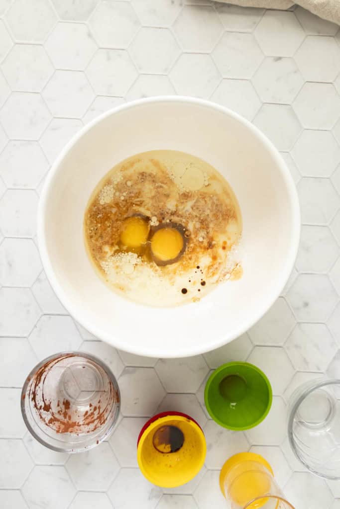 A white mixing bowl on a countertop contains two cracked eggs with additional liquid ingredients. Various empty colored measuring cups and a glass mixing cup surround the bowl.