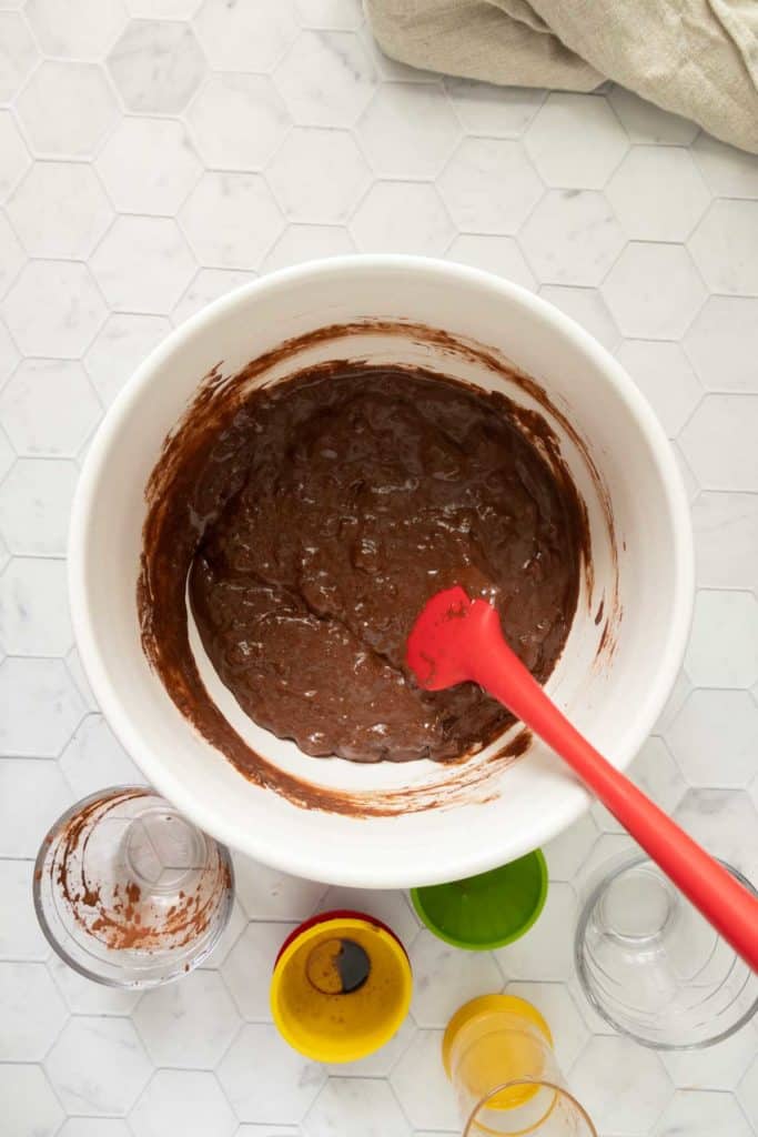 A white mixing bowl with chocolate batter and a red spatula. Several used measuring cups are on the hexagon-tiled surface nearby.