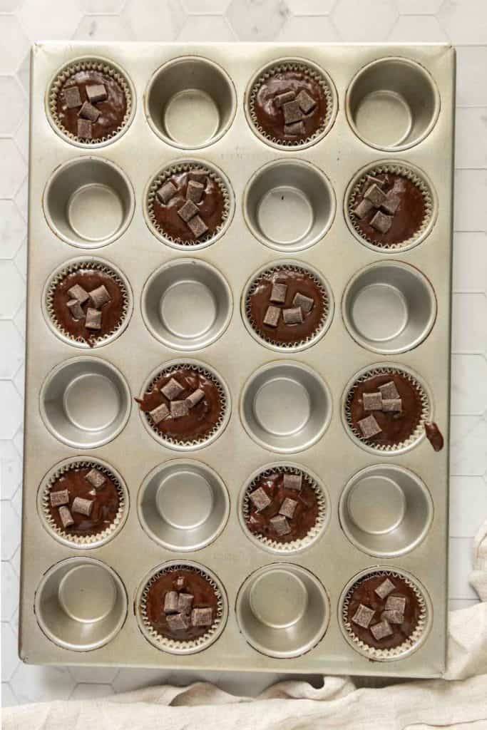 Muffin tin with cups holding chocolate batter topped with chocolate pieces, alongside some empty slots.