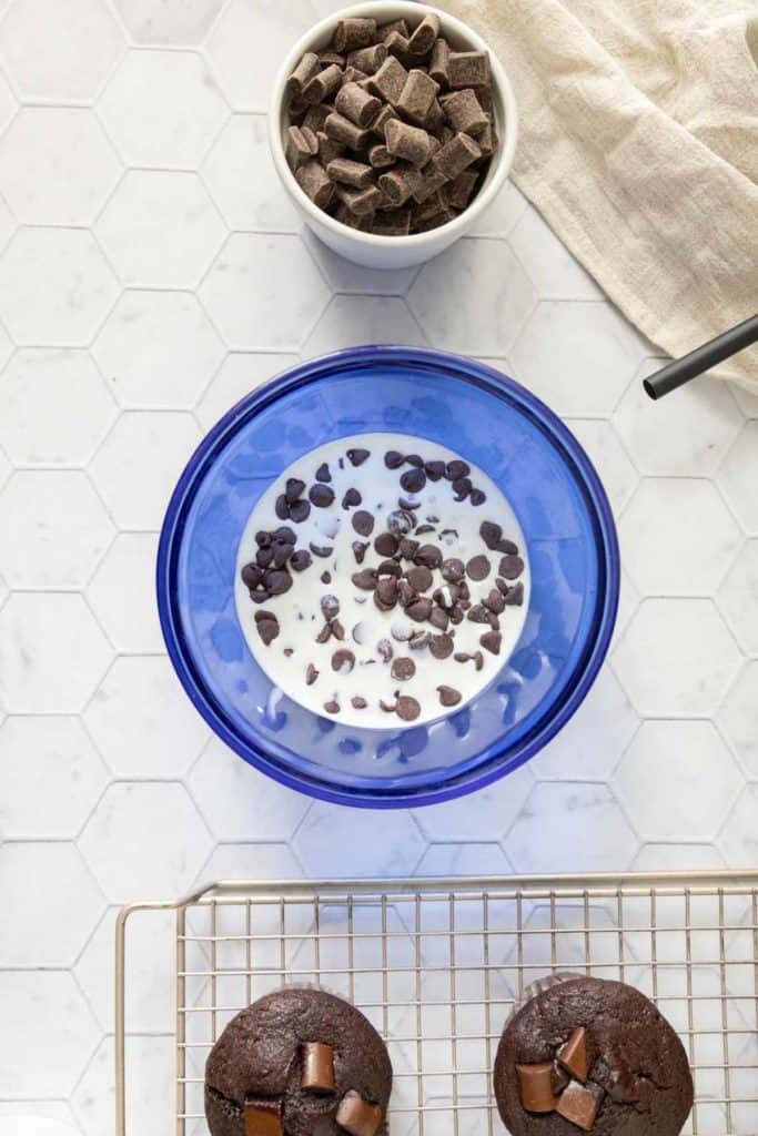 A blue bowl containing milk and chocolate chips is on a hexagonal-tiled countertop next to a small bowl of chocolate chunks. Two chocolate muffins are cooling on a wire rack.