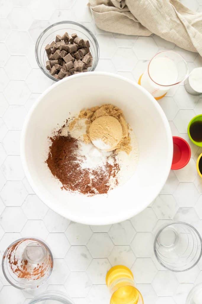 A white mixing bowl containing brown sugar, cocoa powder, and flour on a white countertop, surrounded by glass bowls with chocolate chunks, a cup of milk, red measuring cups, and a beige cloth.