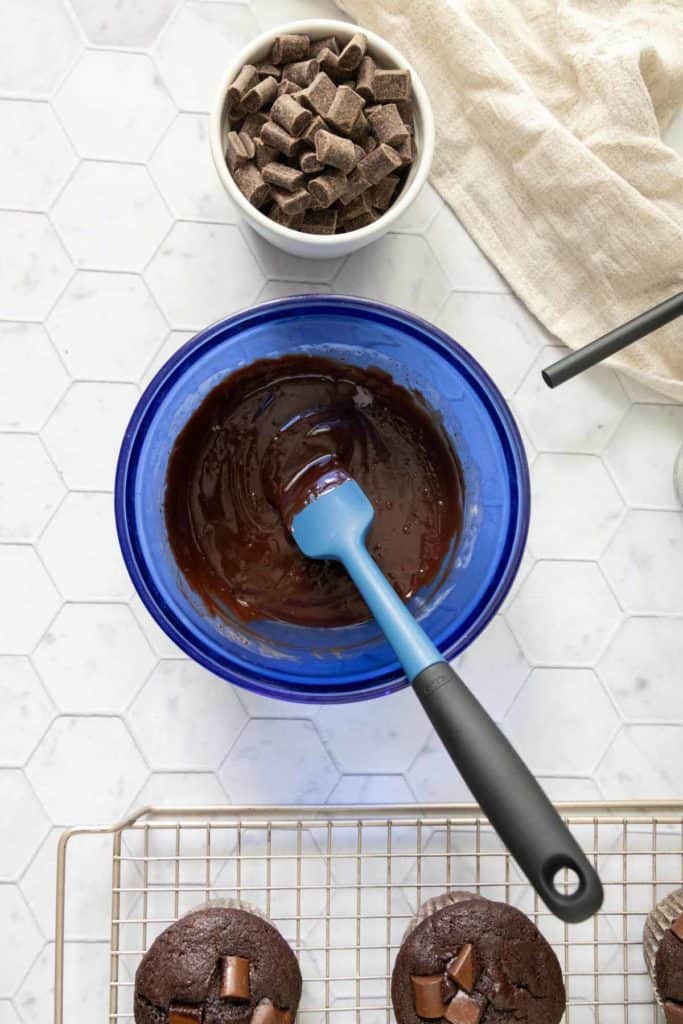 A blue bowl with melted chocolate and a spatula, a small bowl of chocolate chunks, and chocolate muffins on a cooling rack next to a beige cloth on a white hexagon-patterned surface.