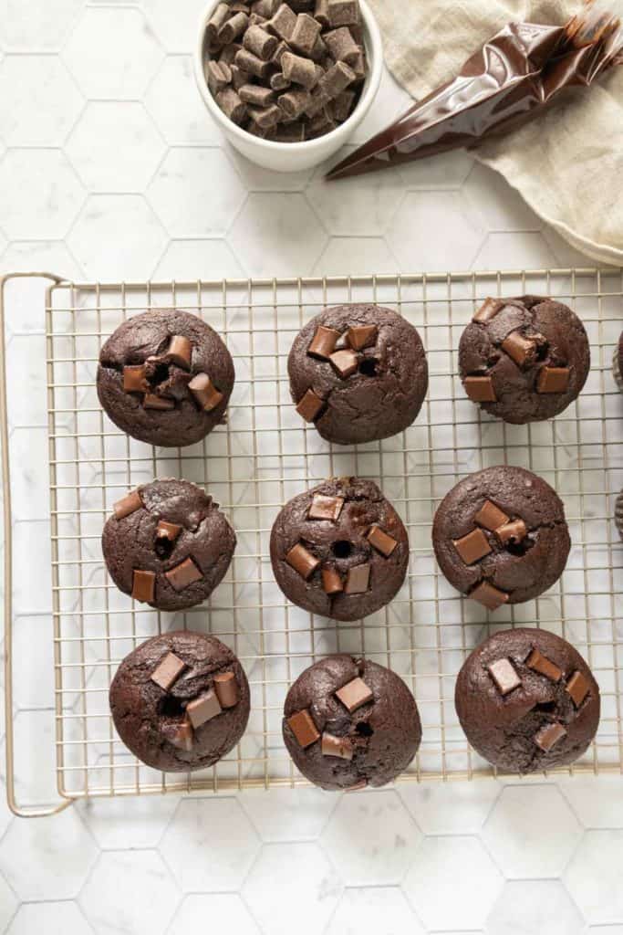 A cooling rack with nine chocolate muffins topped with chocolate chunks. A bowl of chocolate pieces and two piping bags are on the side. The background features a hexagonal tiled surface.