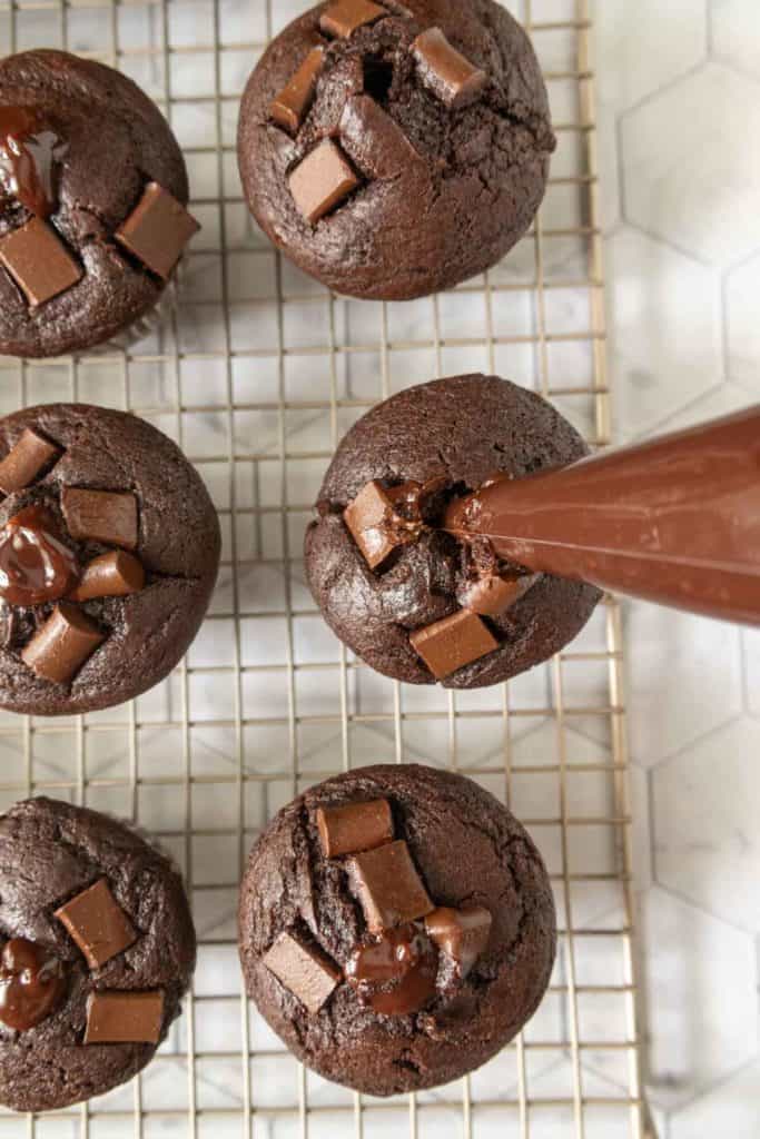 Six chocolate muffins on a cooling rack, some with chocolate chunks on top. A piping bag is adding melted chocolate to the center of one muffin.