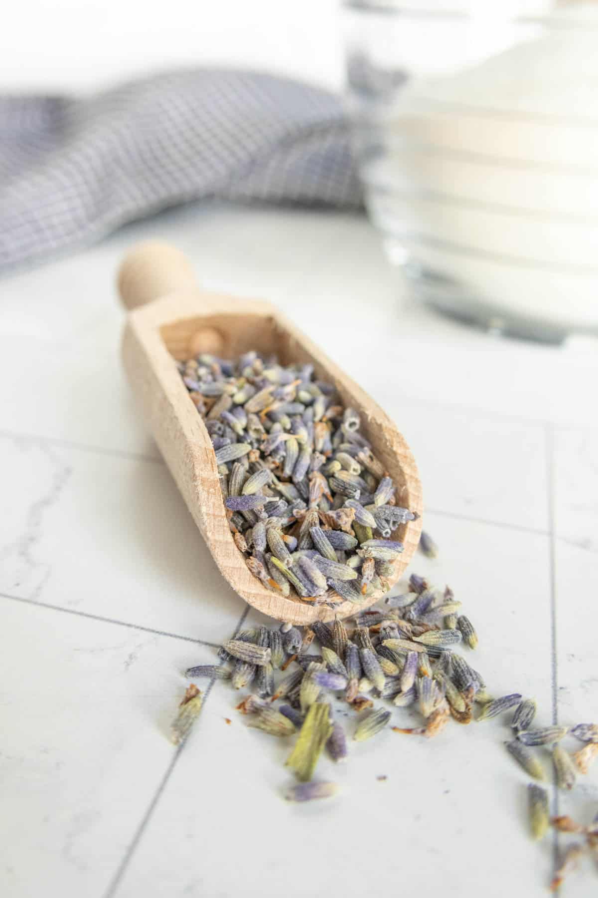 A wooden scoop filled with dried lavender flowers is placed on a marble surface, with a glass jar of white substance and a checkered cloth in the background.