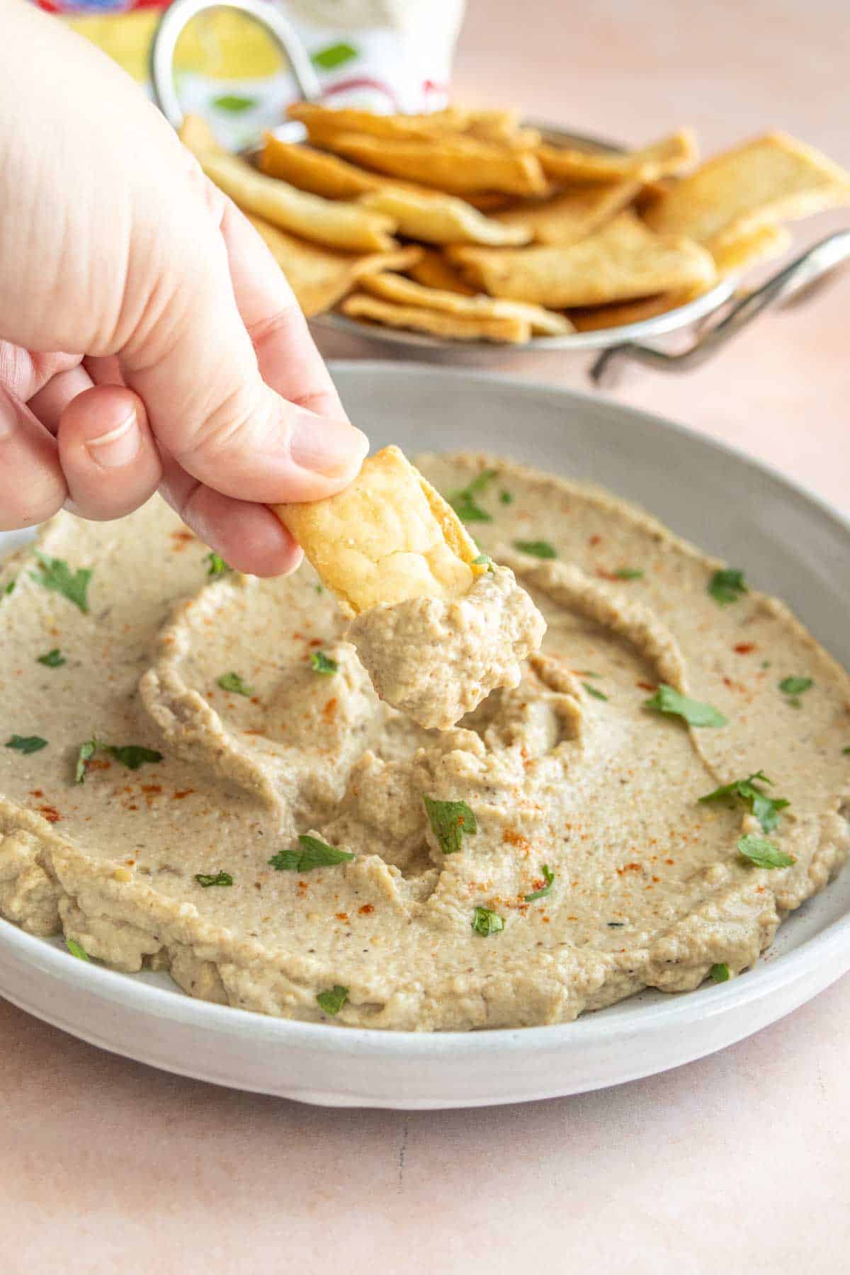 A hand dipping a piece of pita bread into a bowl of smooth eggplant dip, garnished with chopped herbs. A bowl of pita chips is visible in the background.