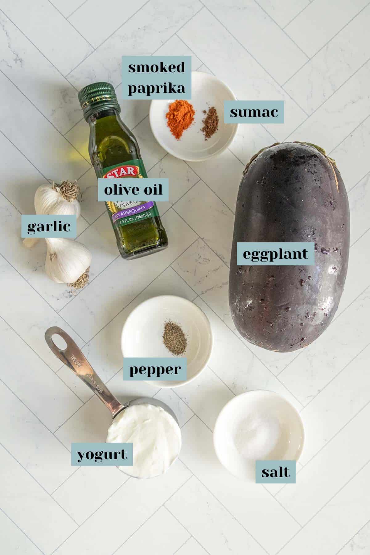 Ingredients for a recipe shown: an eggplant, a bottle of olive oil, two garlic bulbs, and white bowls containing smoked paprika, sumac, pepper, salt, and yogurt.