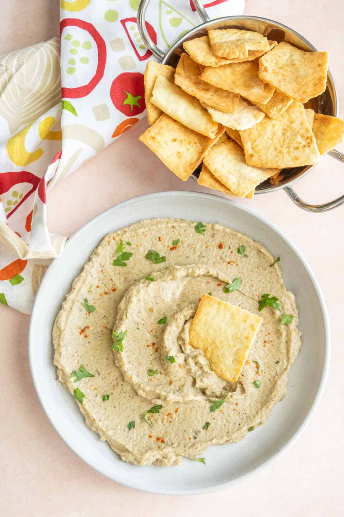 A bowl of eggplant dip garnished with parsley and paprika, accompanied by a metal bowl filled with pita chips. A colorful napkin is partially visible.