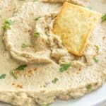 A plate of creamy eggplant dip garnished with chopped herbs and a dash of paprika, topped with a single cracker.