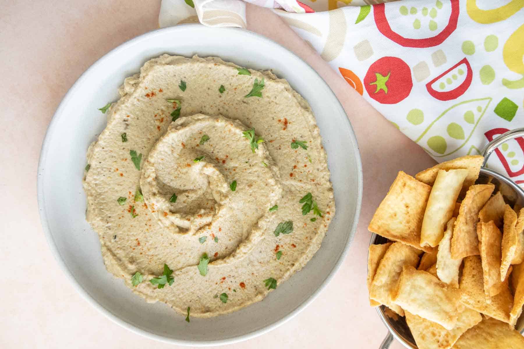 A bowl of eggplant dip garnished with herbs and spices is placed next to a container of pita chips. A colorful fabric with food patterns is partially visible in the background.