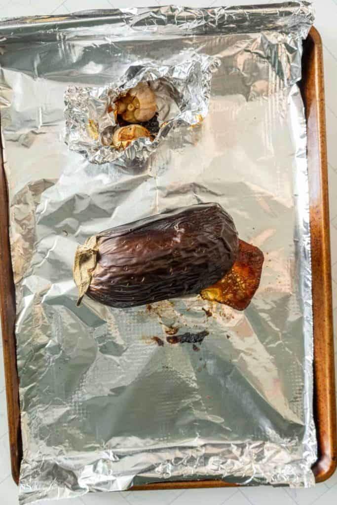 A roasted eggplant and garlic bulb sit on a foil-lined baking sheet.