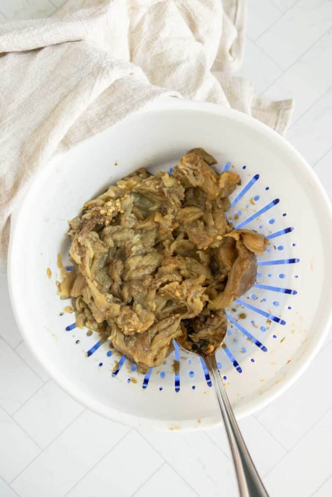 A bowl containing cooked and mashed eggplants with a spoon, positioned on a white tiled surface with a white cloth nearby.