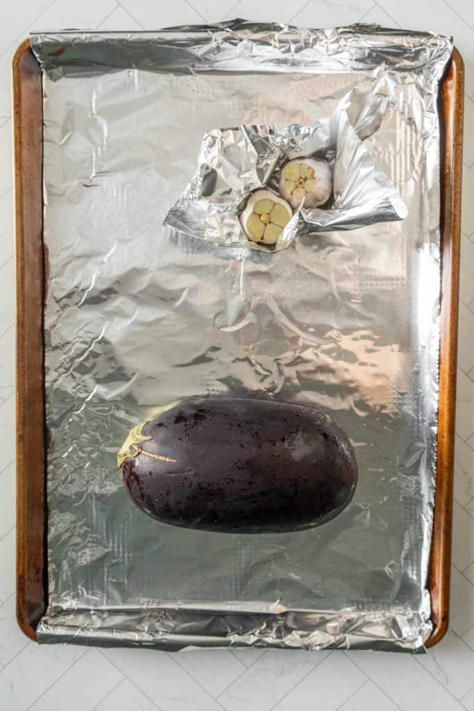 Photo of a foil-lined baking sheet with a whole eggplant and a small foil packet containing halved garlic bulbs on it.