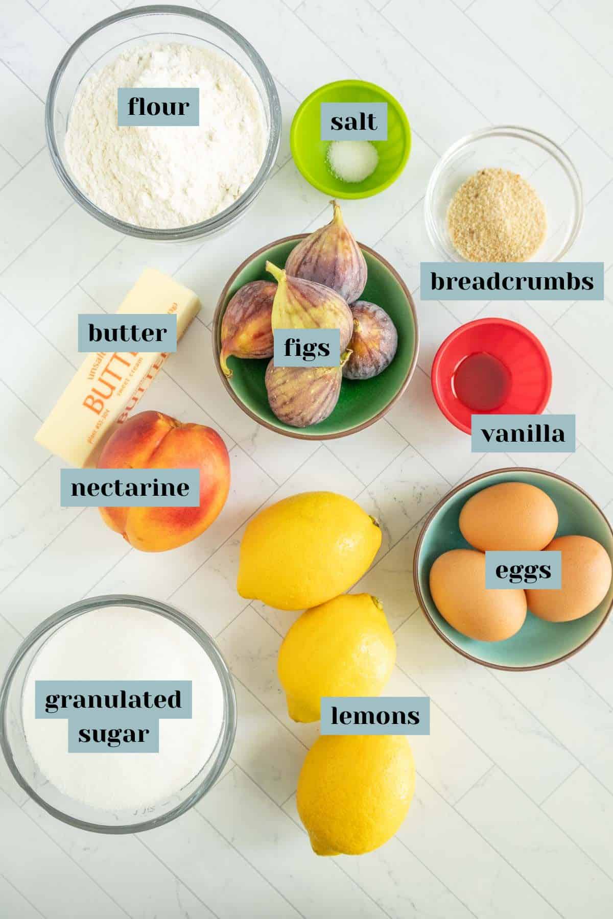 A top view of various labeled food ingredients including flour, salt, breadcrumbs, butter, figs, vanilla, nectarine, eggs, lemons, and granulated sugar on a white countertop.