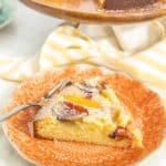 A slice of peach cake with powdered sugar on an orange plate, placed in front of a whole cake on a wooden stand. A fork is on the plate next to the cake slice.