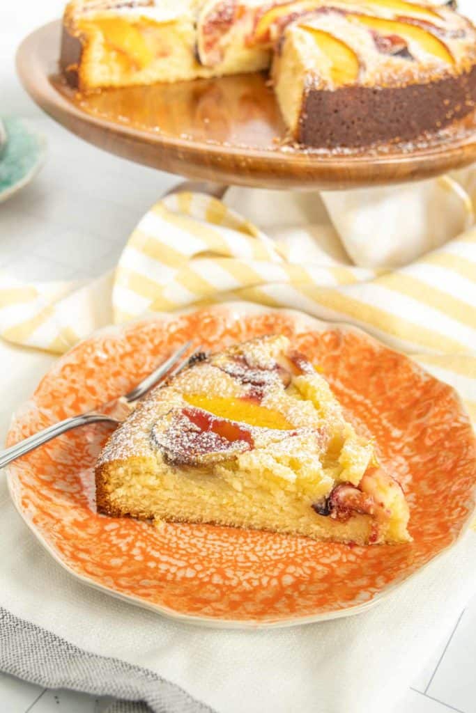 A slice of peach cake with powdered sugar on an orange plate, placed in front of a whole cake on a wooden stand. A fork is on the plate next to the cake slice.